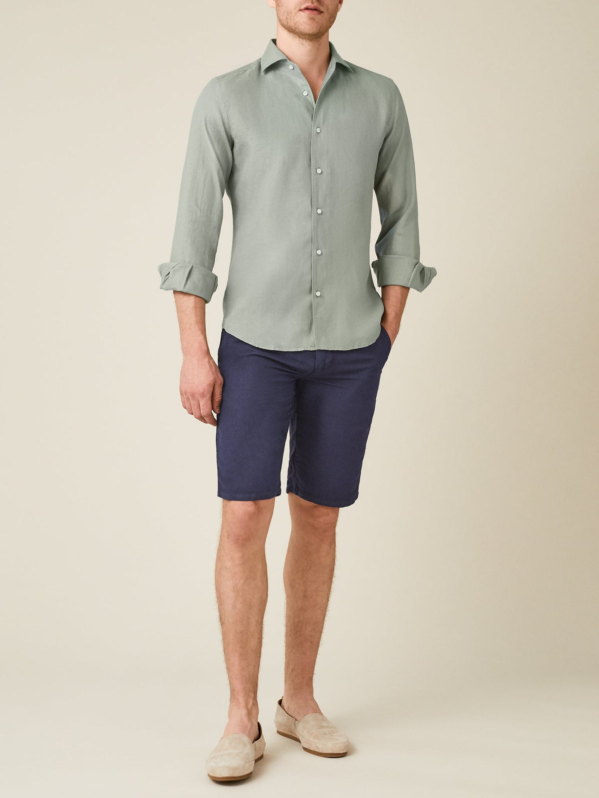 A man models the Portofino Linen Shirt in Sage Green, styled with navy shorts and loafers against a plain background.