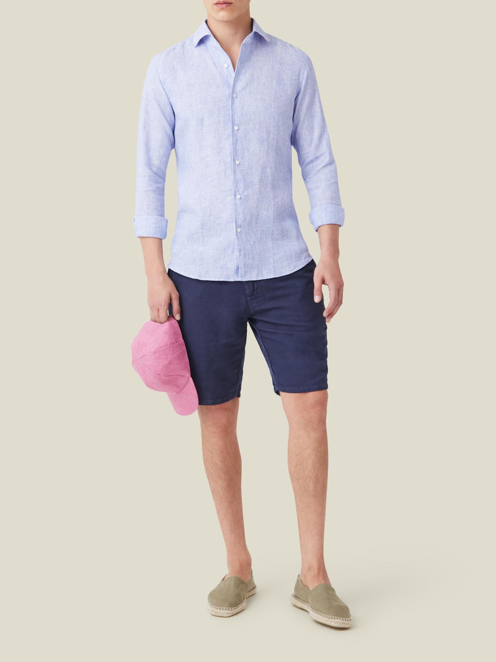Model wears Striped Classic Blue Portofino Linen Shirt, navy shorts, and espadrilles, holding a pink cap.