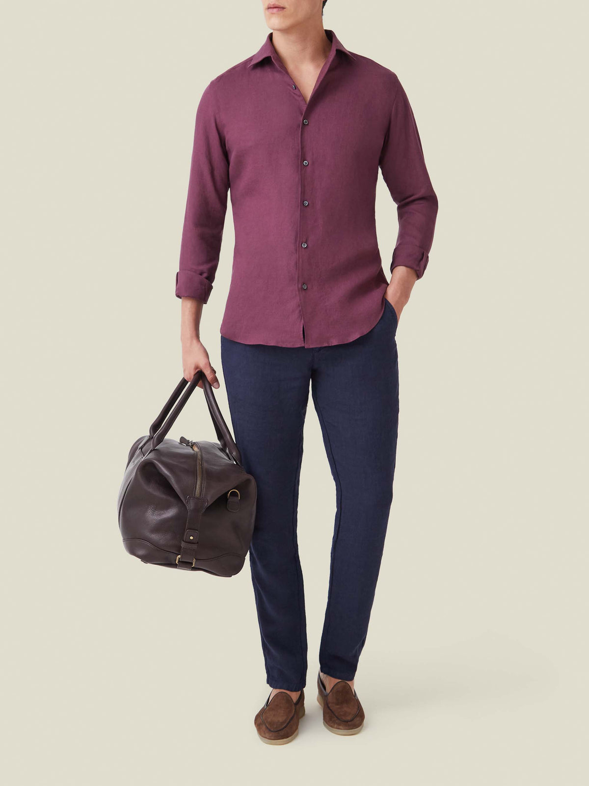A model wears the Portofino Linen Shirt in Burgundy, styled with navy trousers, brown loafers, and a leather travel bag.