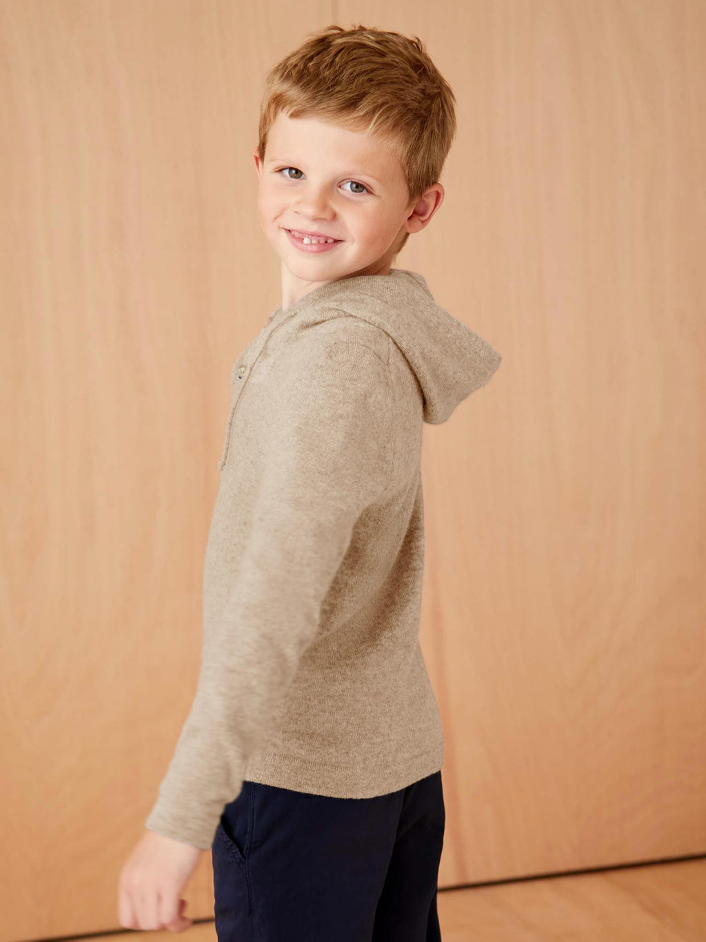 Side profile of a young boy smiling while wearing the Luca Faloni Junior Cashmere Hoodie in Camel Beige.