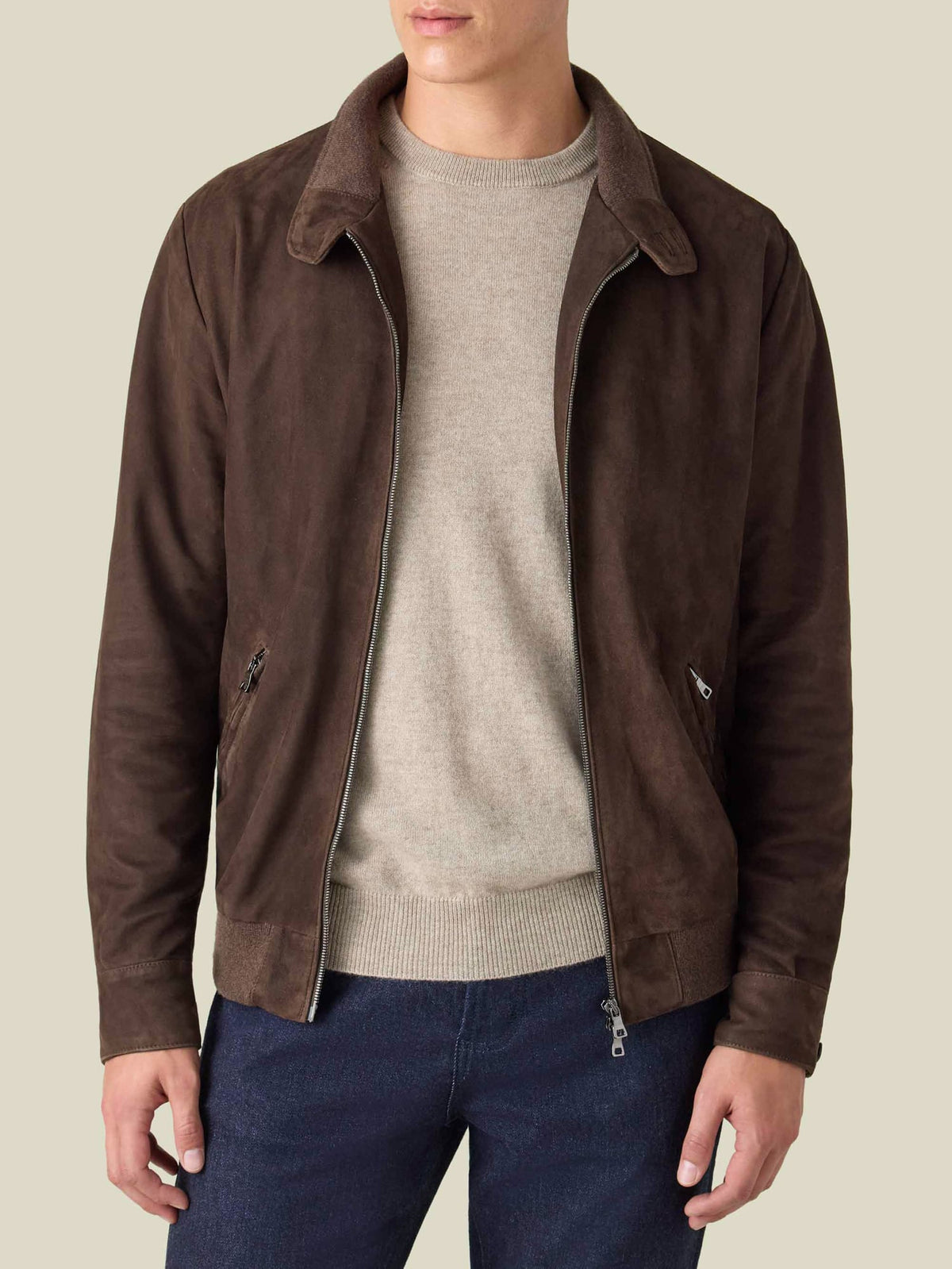 A model wears the Classic Bomber Jacket in chocolate brown goatskin suede, open over a beige sweater and dark jeans.