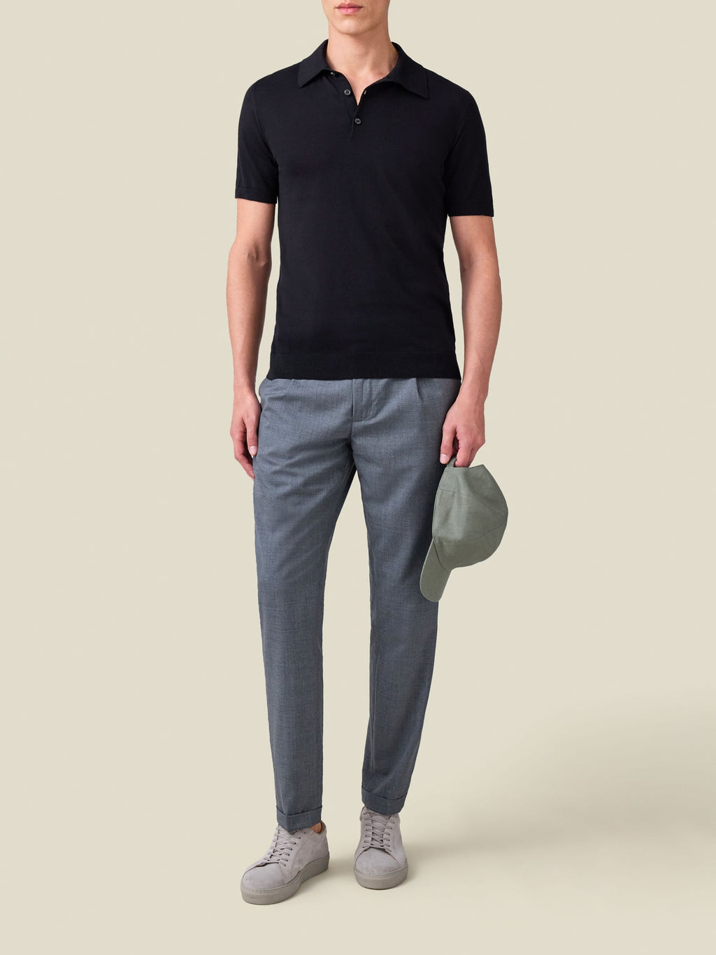 A model wears the black Knitted Cotton Polo with grey trousers and light grey sneakers, holding a green cap.