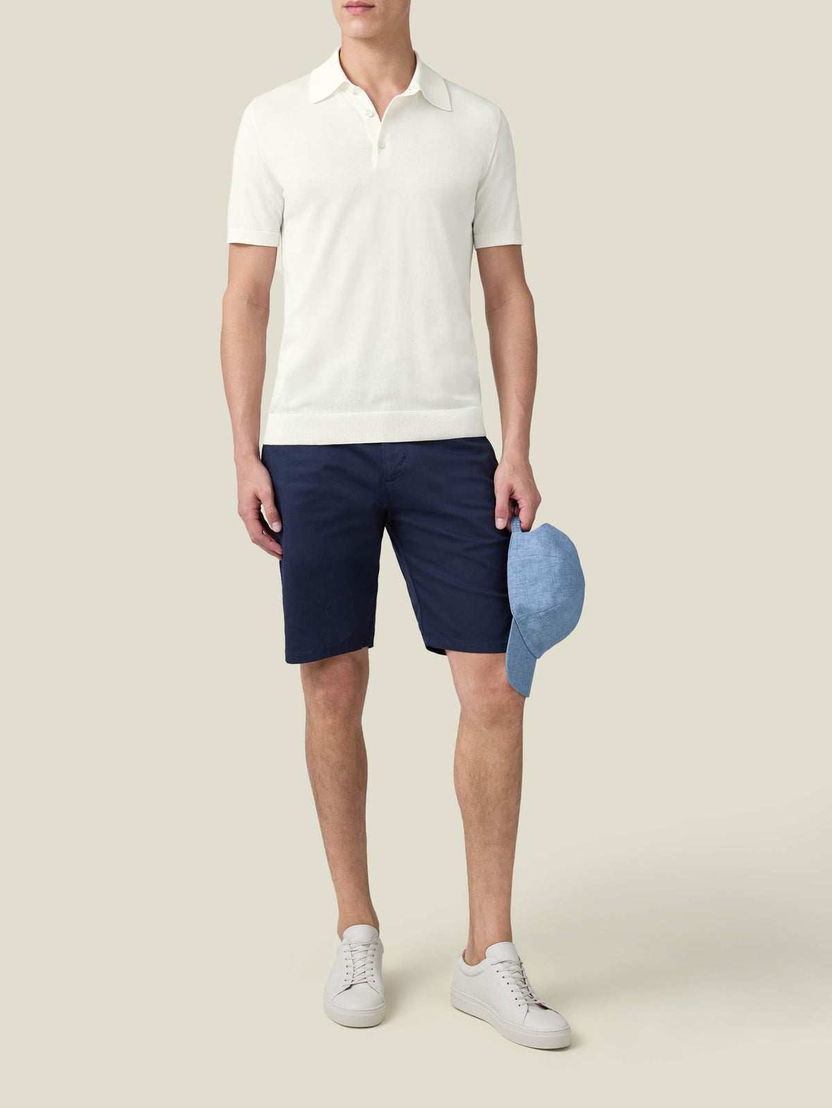 A front view of a model wearing the Knitted Cotton Polo in cream, with navy shorts, white trainers and a blue cap.