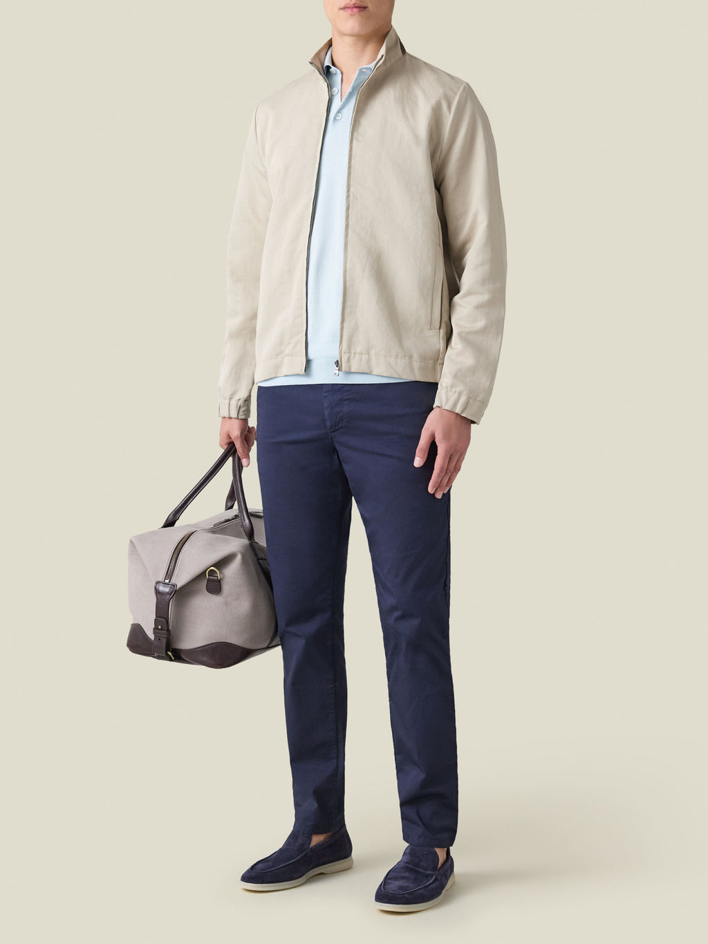 Man wearing the light blue Knitted Cotton Polo, styled with a beige zip jacket, navy chinos, and holding a duffel bag.