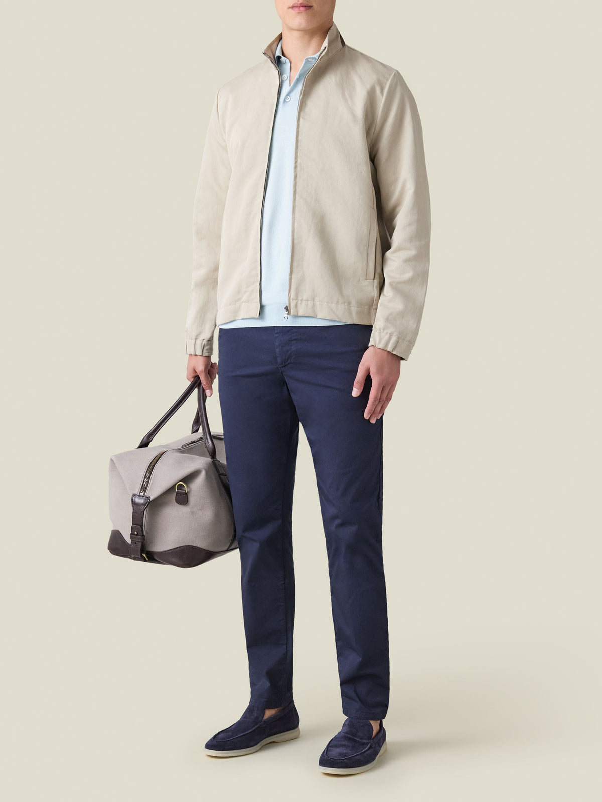Man wearing the light blue Knitted Cotton Polo, styled with a beige zip jacket, navy chinos, and holding a duffel bag.