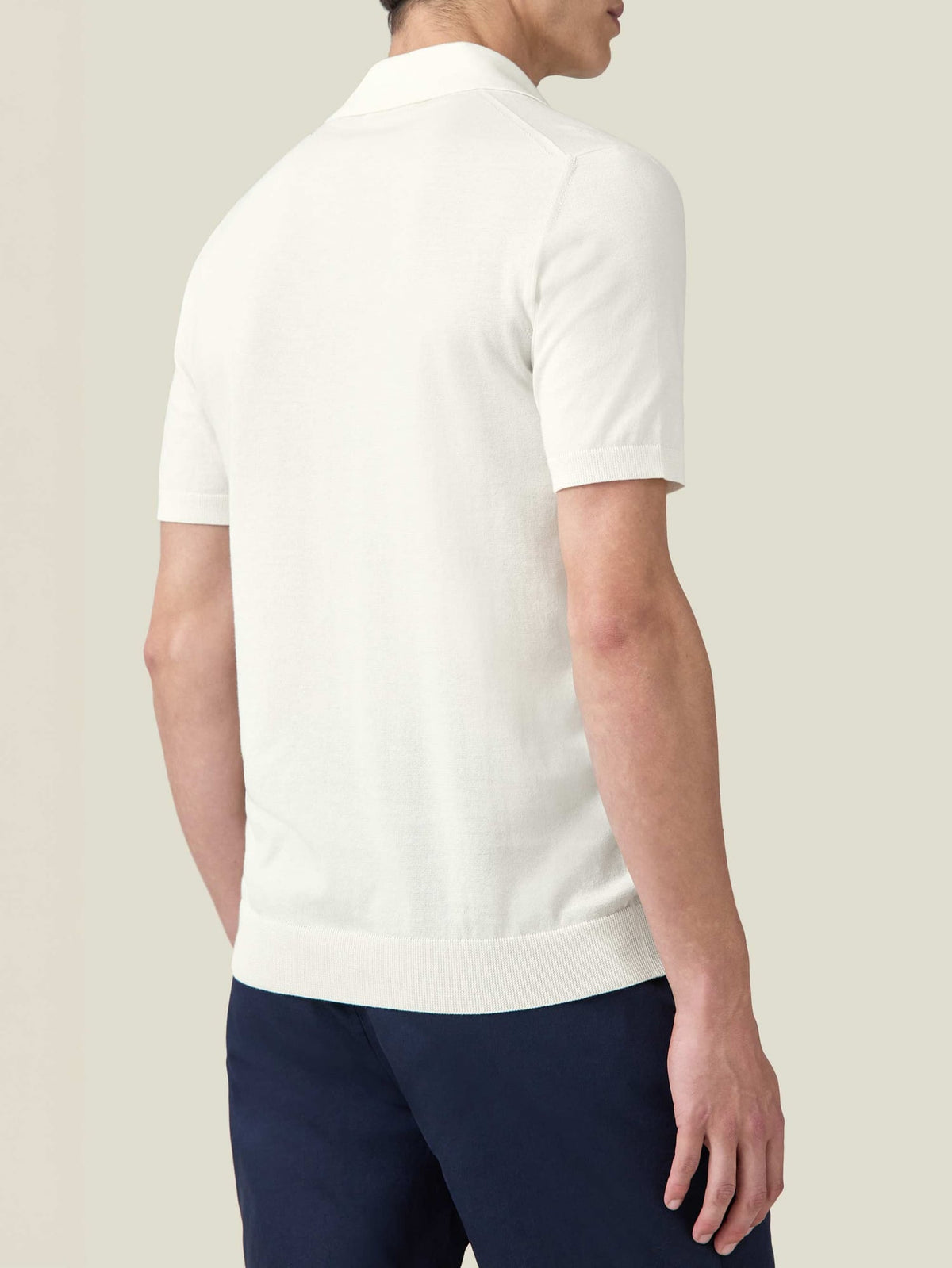 A rear three-quarter view of a model wearing the Luca Faloni cream Knitted Cotton Polo with navy trousers.