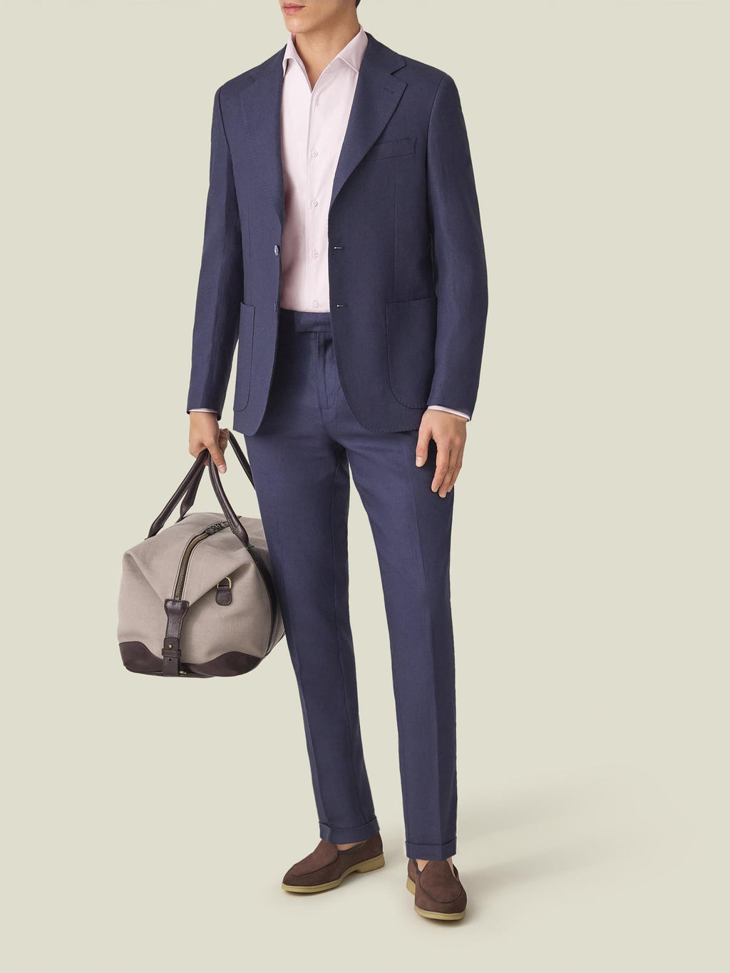 A model wears the light pink Lightweight Poplin Cotton Shirt with a navy suit, brown loafers, and a grey travel bag.