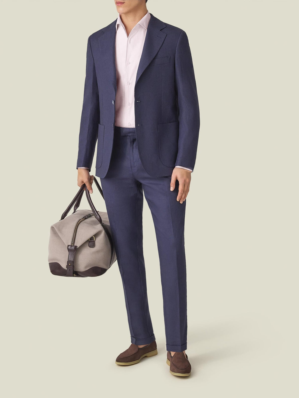 A model wears the light pink Lightweight Poplin Cotton Shirt with a navy suit, brown loafers, and a grey travel bag.
