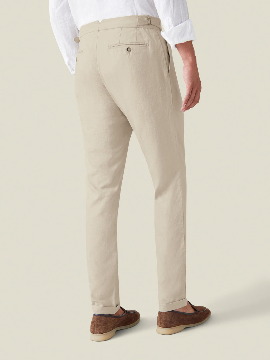 A man from behind wearing slim-fit sand linen trousers with cuffed ankles, paired with a white shirt and brown suede loafers.