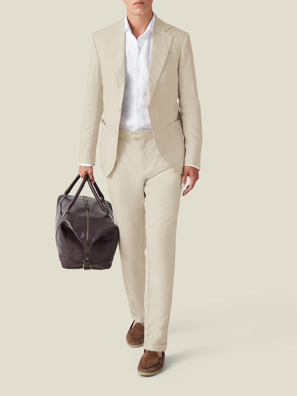 A model wears the Sand Linen Elegant Trousers and matching blazer, styled with a white shirt and a brown leather travel bag.