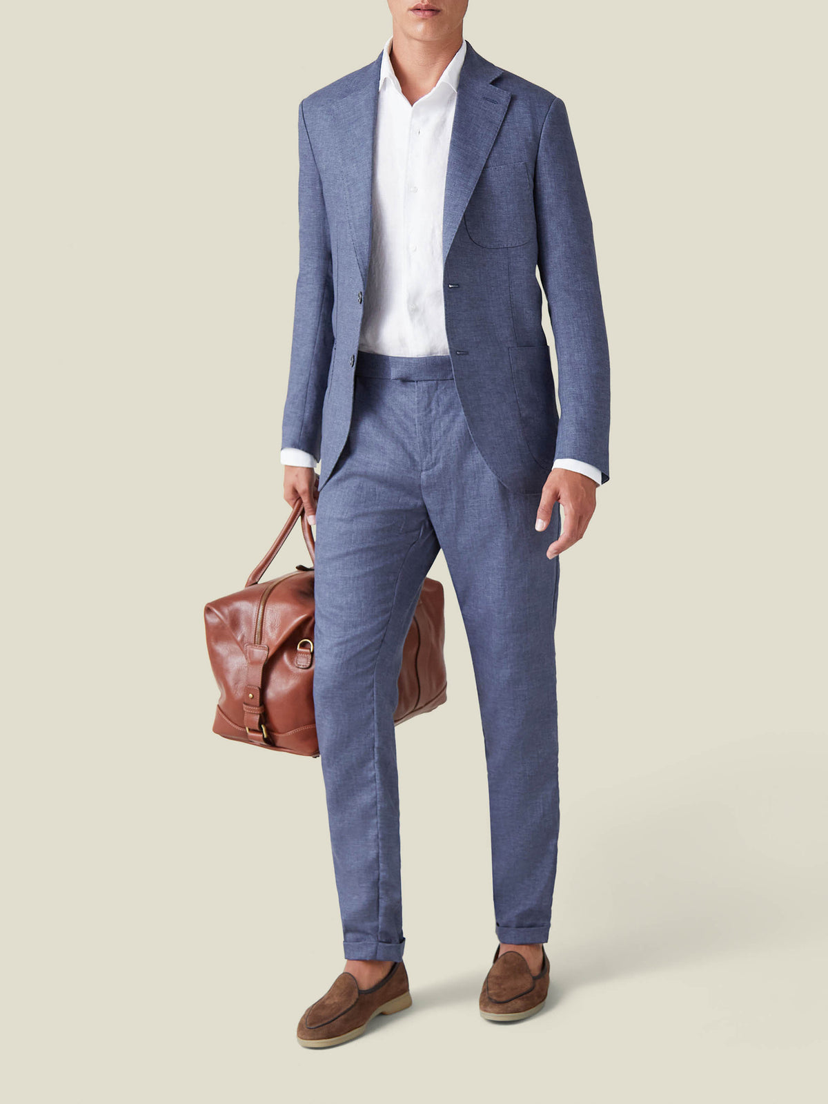 A model wears the Luca Faloni Linen Suit in Azure, styled with a white shirt, brown loafers, and a brown leather bag.