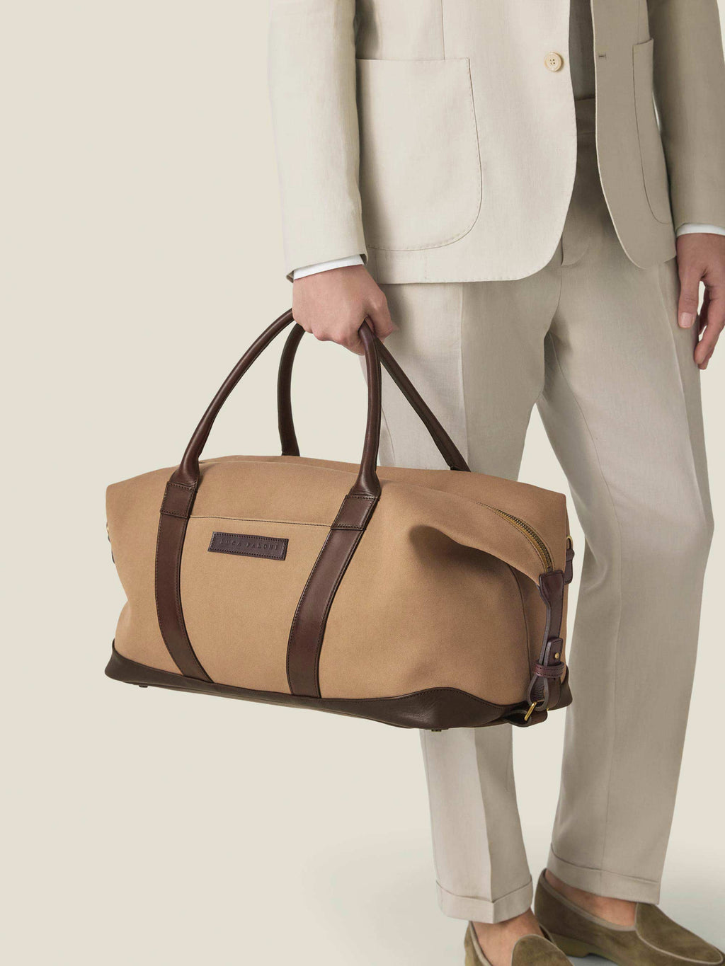 Model wearing a light beige suit holds the Luca Faloni Canvas Weekender Bag in Caramel Brown by its dark leather handles.