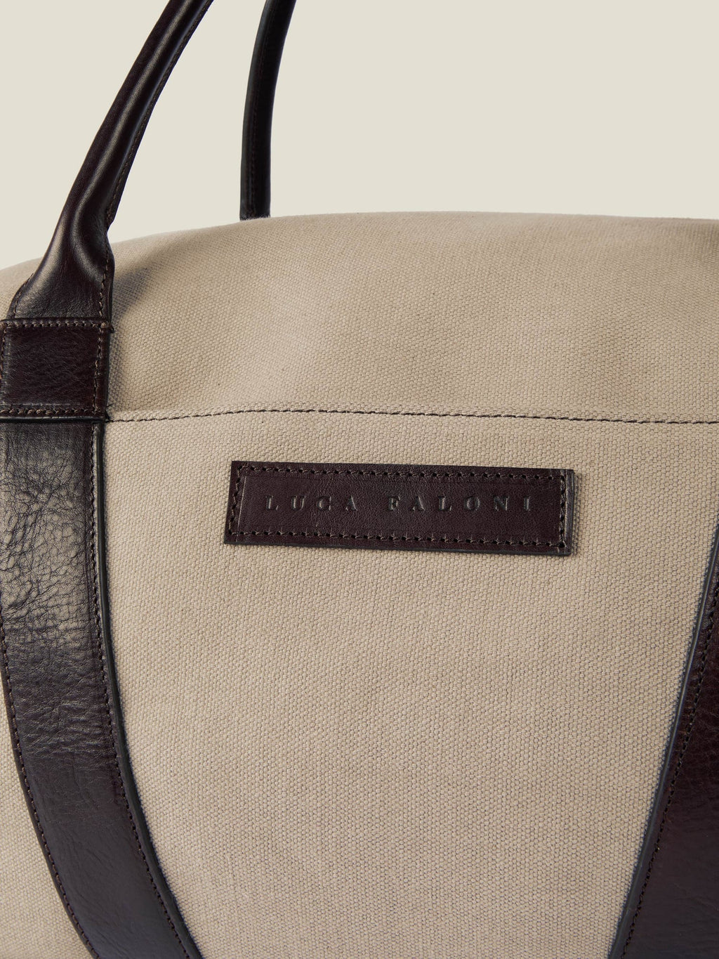 Close-up of the Luca Faloni Canvas Bag in sand, showing the embossed leather logo patch and dark brown leather handle.