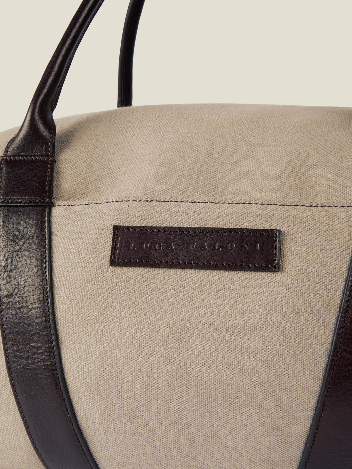 Close-up of the Luca Faloni Canvas Bag in sand, showing the embossed leather logo patch and dark brown leather handle.