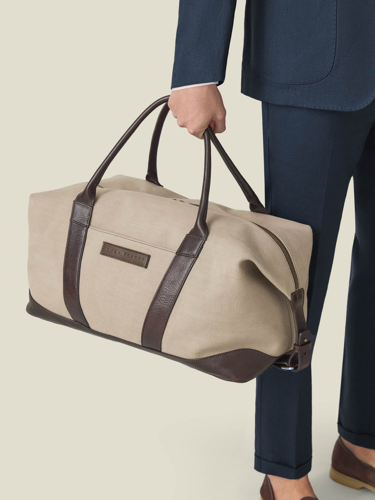 Close-up of the Luca Faloni Sand Canvas Bag with brown leather trim, carried by a man in a navy suit.